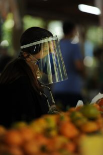 A foto mostra uma pessoa usando viseira facial transparente e máscara de proteção, em ambiente que parece ser uma feira. Em primeiro plano, e em desfoque há uma bancada com frutas e o fundo, também desfocado, indica movimento e presença de outras pessoas.