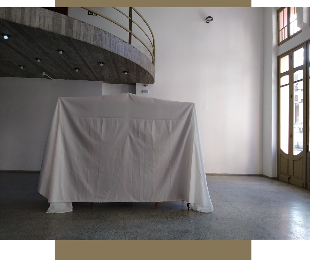 Imagem colorida. Em ambiente interno de galeria expositiva, ao centro da foto, enquadra-se um guarda-roupas coberto por um tecido branco.