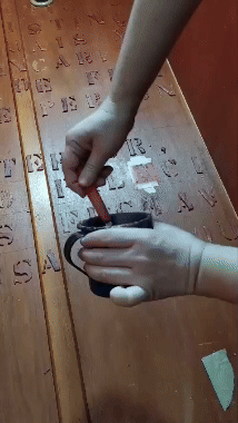  Imagem colorida. Enquadra uma pessoa manipulando uma substância líquida e a depositando em letras em alto relevo no fundo interior de um guarda-roupas