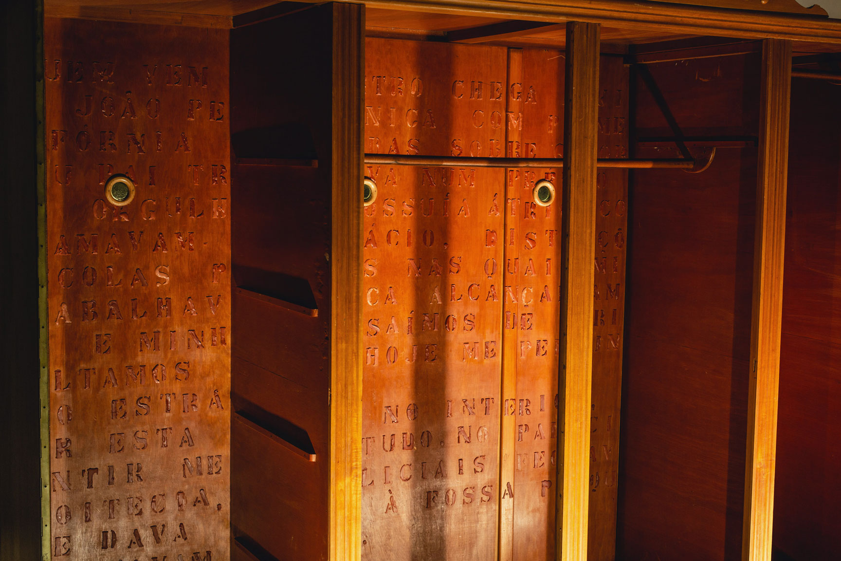 Imagem colorida. Em plano único, recorte de um guarda-roupa em cerejeira, sem portas, em um espaço expositivo, é iluminado. Na parte interior do objeto consta o seguinte texto: “Quem vem do centro chega aqui pela Ponte da João Pessoa, única com Palmeiras-da-Califórnia plantadas sobre o dorso. Seguindo uma quadra adiante, no sentido do rio, encontra-se a Ponte da Azenha. Suas tochas de pedra são uma cortesia dos antigos para iluminar o trajeto daqueles que saiam da cidade em direção ao camposanto. Meu pai trabalhava num prédio do qual eu me orgulhava. Possuía átrio, colunas e chamavam-no Palácio, distinções que as escolas públicas nas quais minha mãe trabalhou nunca alcançariam. Eu e minha mãe saímos de férias e não retornamos. Hoje me pergunto por que, na época, não estranhei. Voltamos uma única manhã quando a casa não funcionava mais. Abrimos a dispensa e cozinhamos macarrão para comer de café. Por aquela época, no interior, comecei a desenvolver medo patológico. No Palácio, quando anoitecia, os policiais fechavam a porta que dava acesso à fossa porque ainda escutavam gritos.”