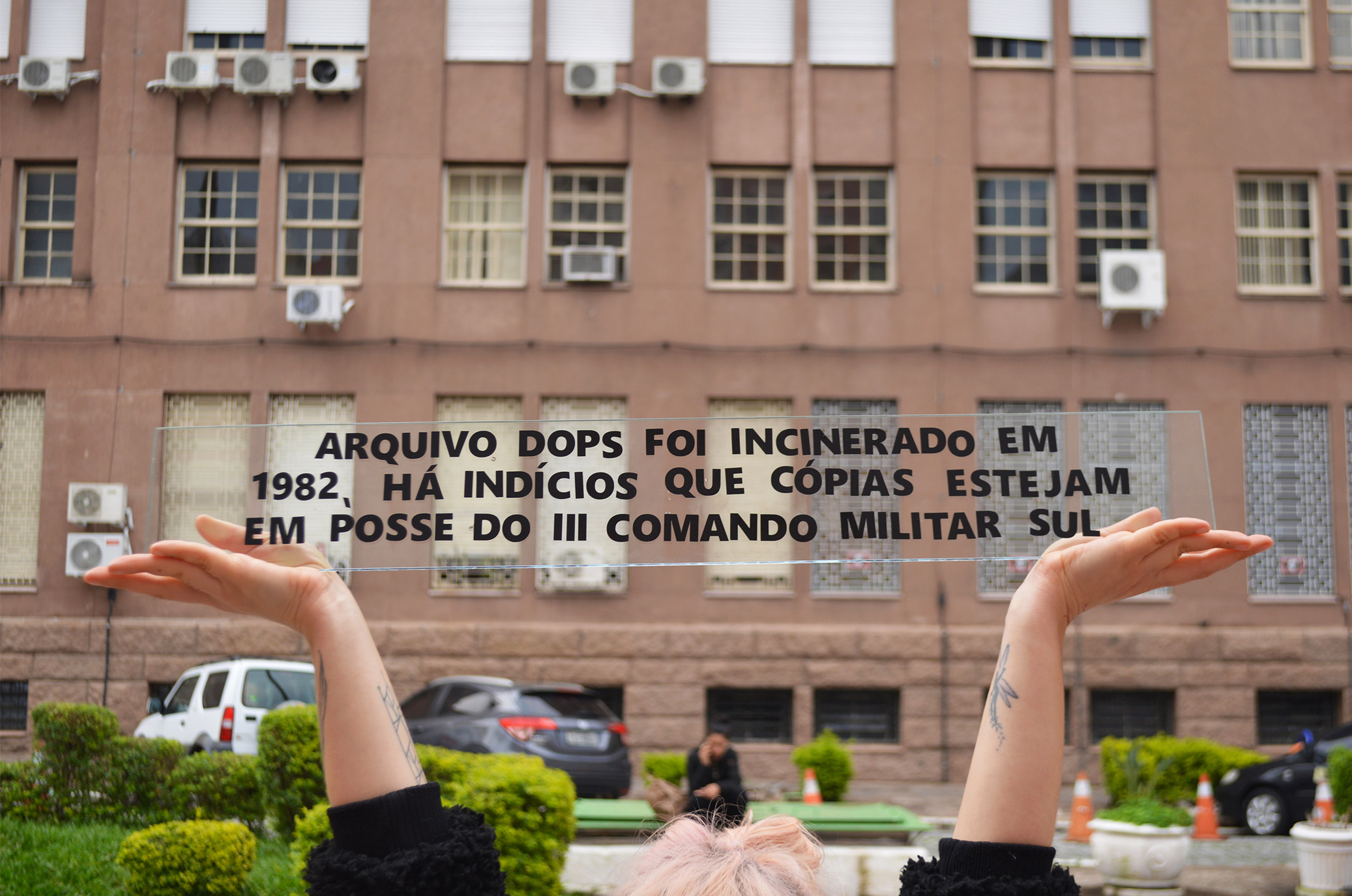 Fotografia colorida. Em primeiro plano uma pessoa carrega uma placa acrílica transparente com a seguinte mensagem: Arquivo DOPS foi incinerado em 1982, há indícios que cópias estejam em posse do III Comando Militar Sul. Ao fundo enquadra-se a fachada dessa instituição.