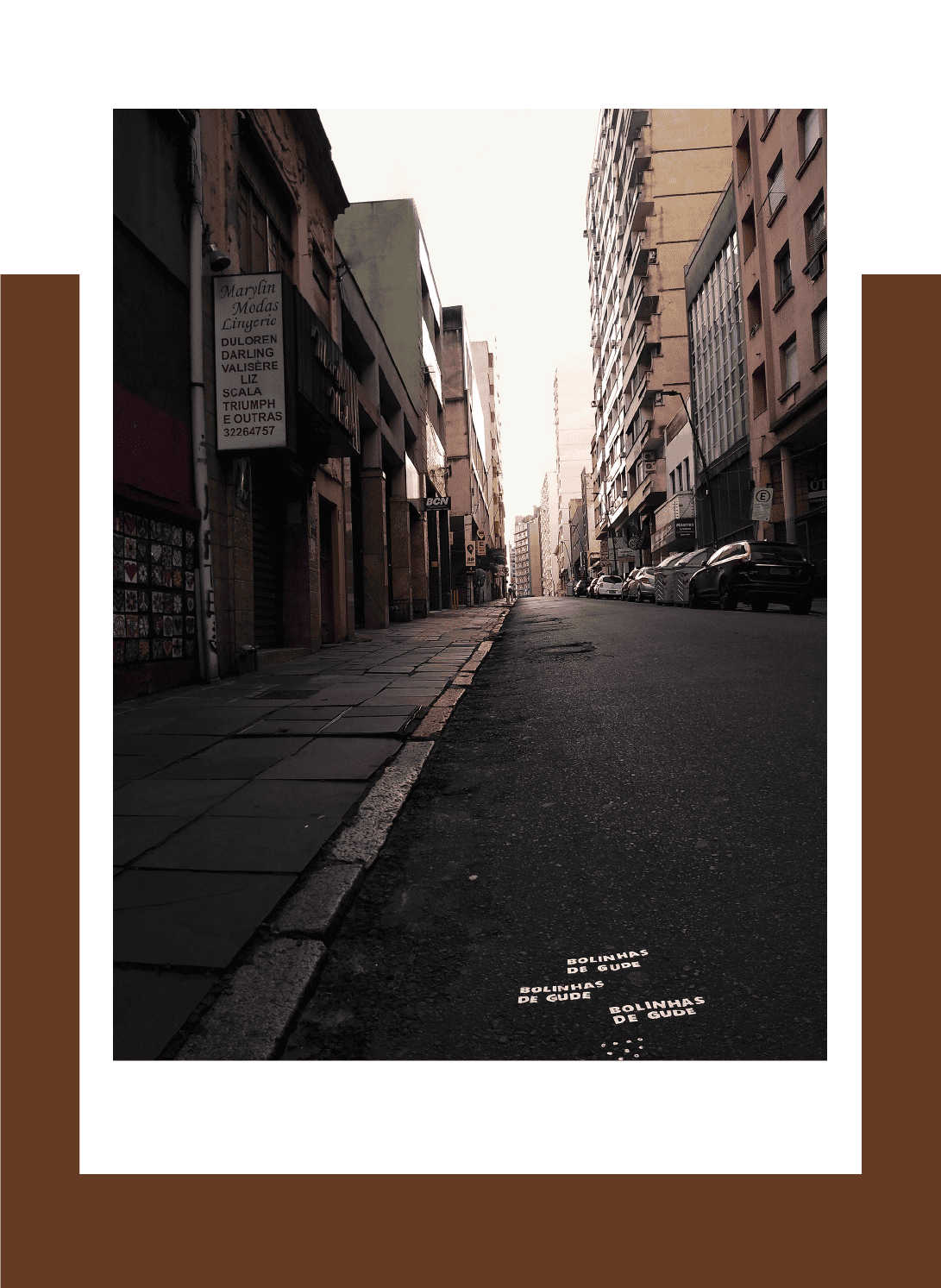 Fotografia colorida. Em plano central enquadra a via pública. No asfalto foi plasmada a seguinte mensagem: Bolinhas de gude.. Bolinhas de gude… Bolinhas de gude. Nos planos laterais estão alguns edifícios da Rua Riachuelo em Porto Alegre.