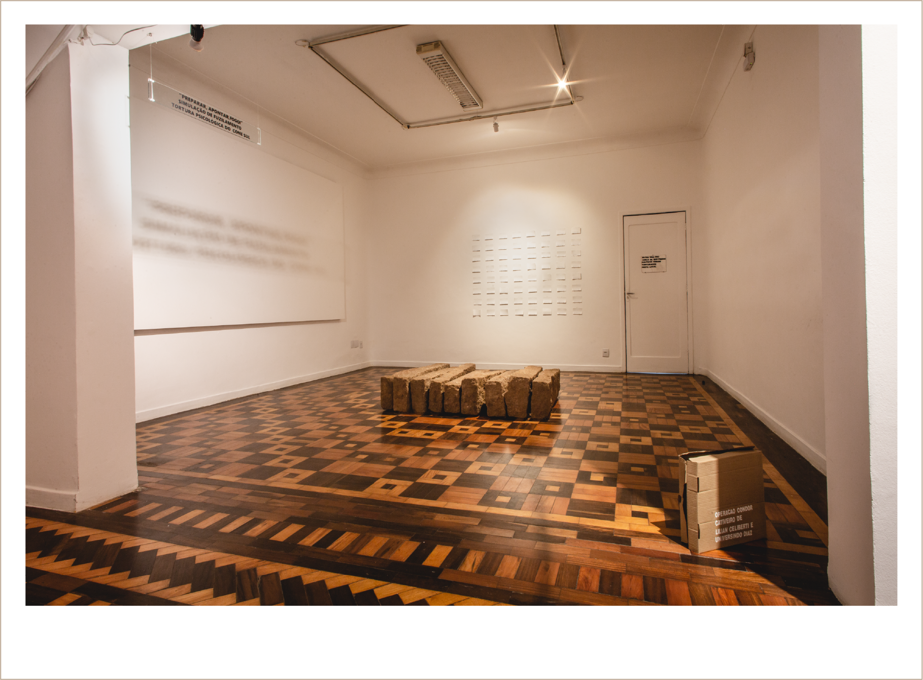 Imagem colorida. Em plano aberto retrata espaço expositivo. No piso, repousa uma caixa de papelão com colagem que carrega a seguinte mensagem: Operação Condor Cativeiro de Lilian Celiberti e Universindo Diaz. Ao centro da galeria, repousam no chão nove blocos de meios-fios de granito.