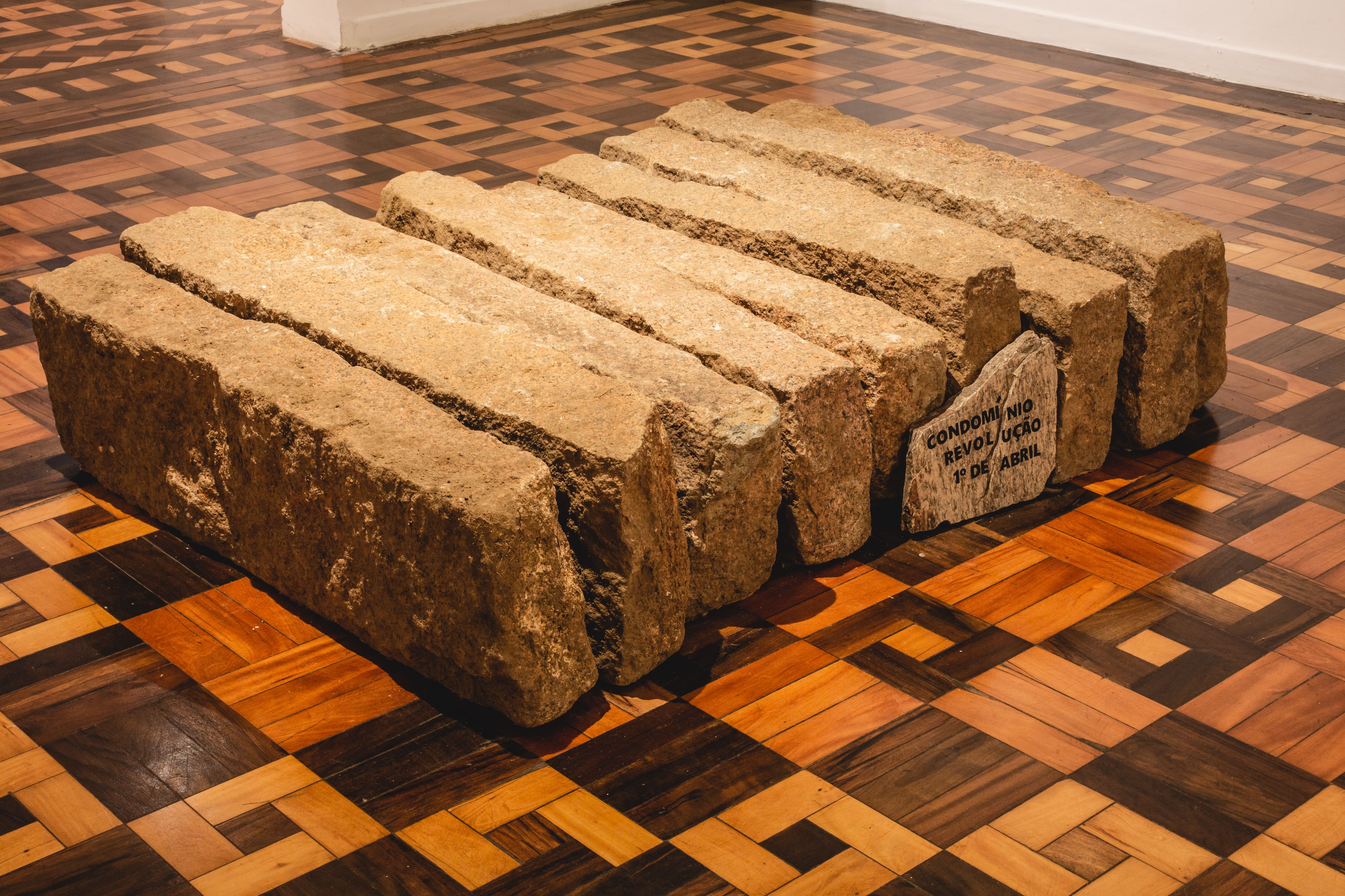 Fotografia colorida. Em plano central estão repousados sob o piso de uma galeria 9 blocos de meios-fios de granito. Apoiado em um deles um fragmento da rocha carrega a seguinte mensagem: Condomínio Revolução Primeiro de Abril.