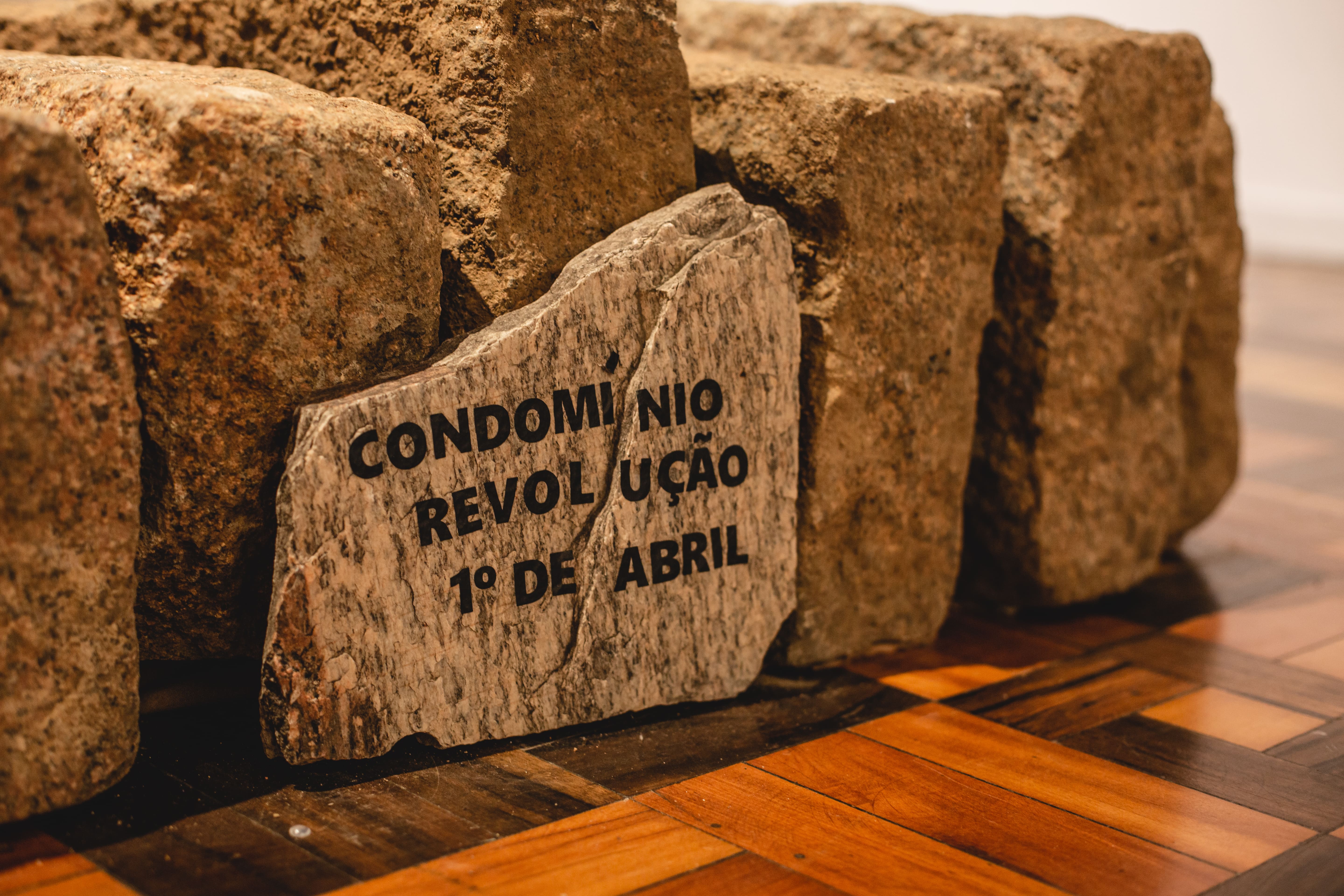 Fotografia colorida. Estão retratados blocos de meios-fios de granito repousados sob o piso de uma galeria. Apoiado em um deles um fragmento da rocha carrega a seguinte mensagem: Condomínio Revolução Primeiro de Abril.