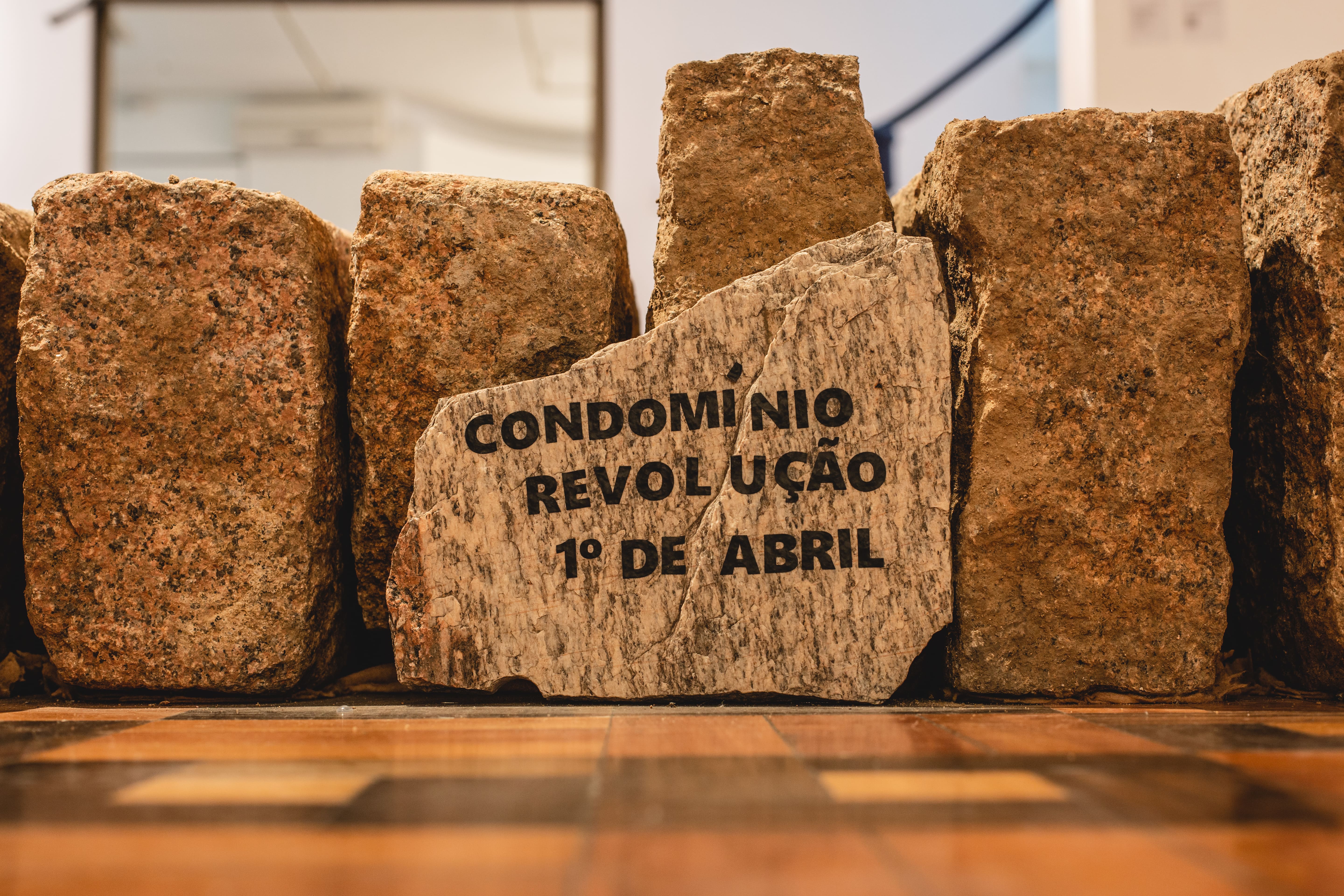 Fotografia colorida. Estão retratados blocos de meios-fios de granito repousados sob o piso de uma galeria. Apoiado em um deles um fragmento da rocha carrega a seguinte mensagem: Condomínio Revolução Primeiro de Abril. 