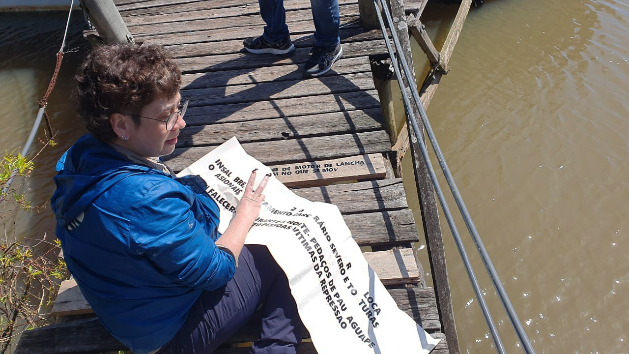 Fotografia colorida. Em plano único, Manoela está sentada em uma passarela de madeira sob a água. Ela carrega material dos epigramas, enquanto decalca letra por letra na superfície de madeira