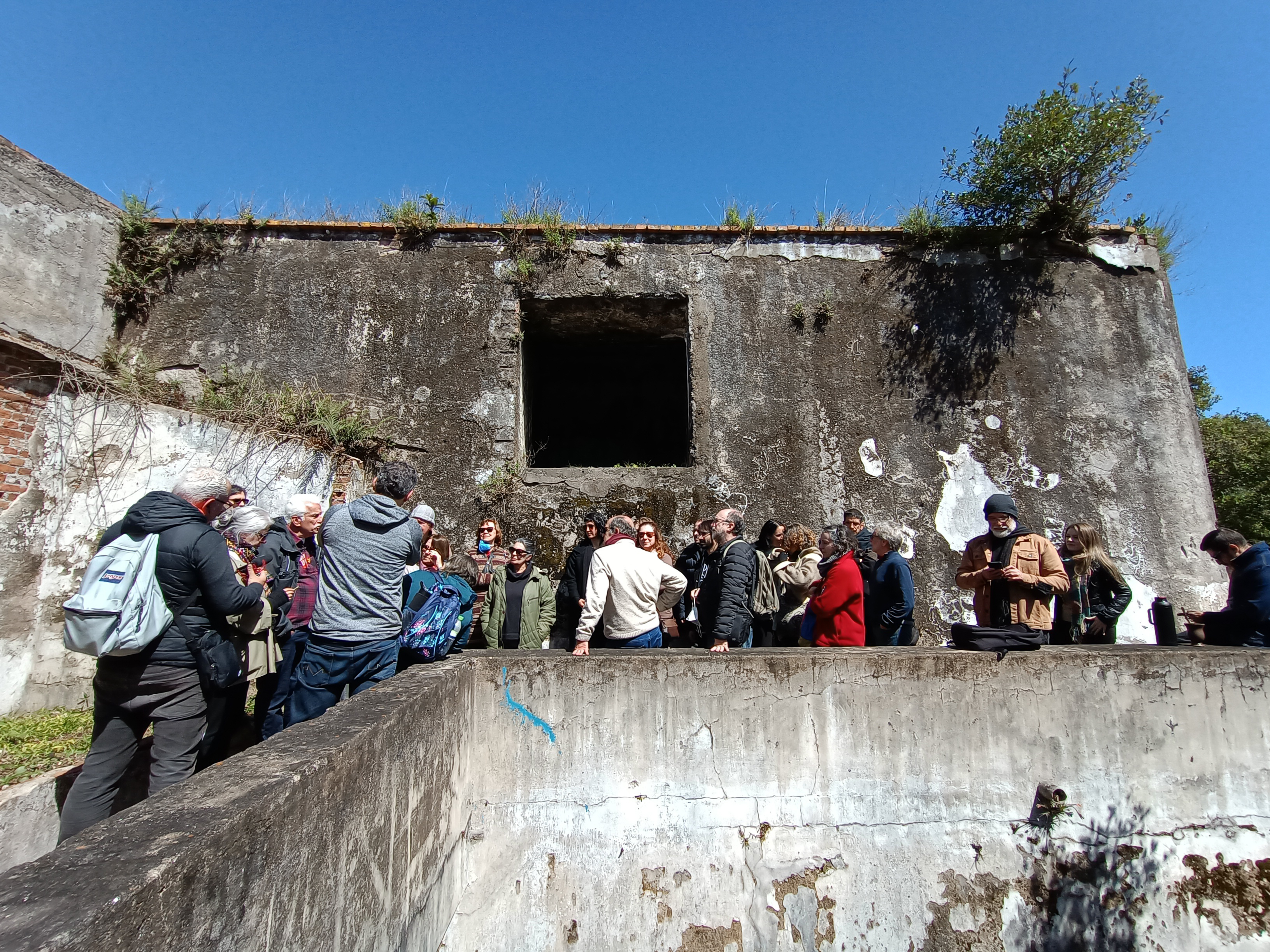 Imagem colorida. Enquadra um grupo de aproximadamente 20 pessoas transitando em área externa de ruínas na Ilha do Presídio.