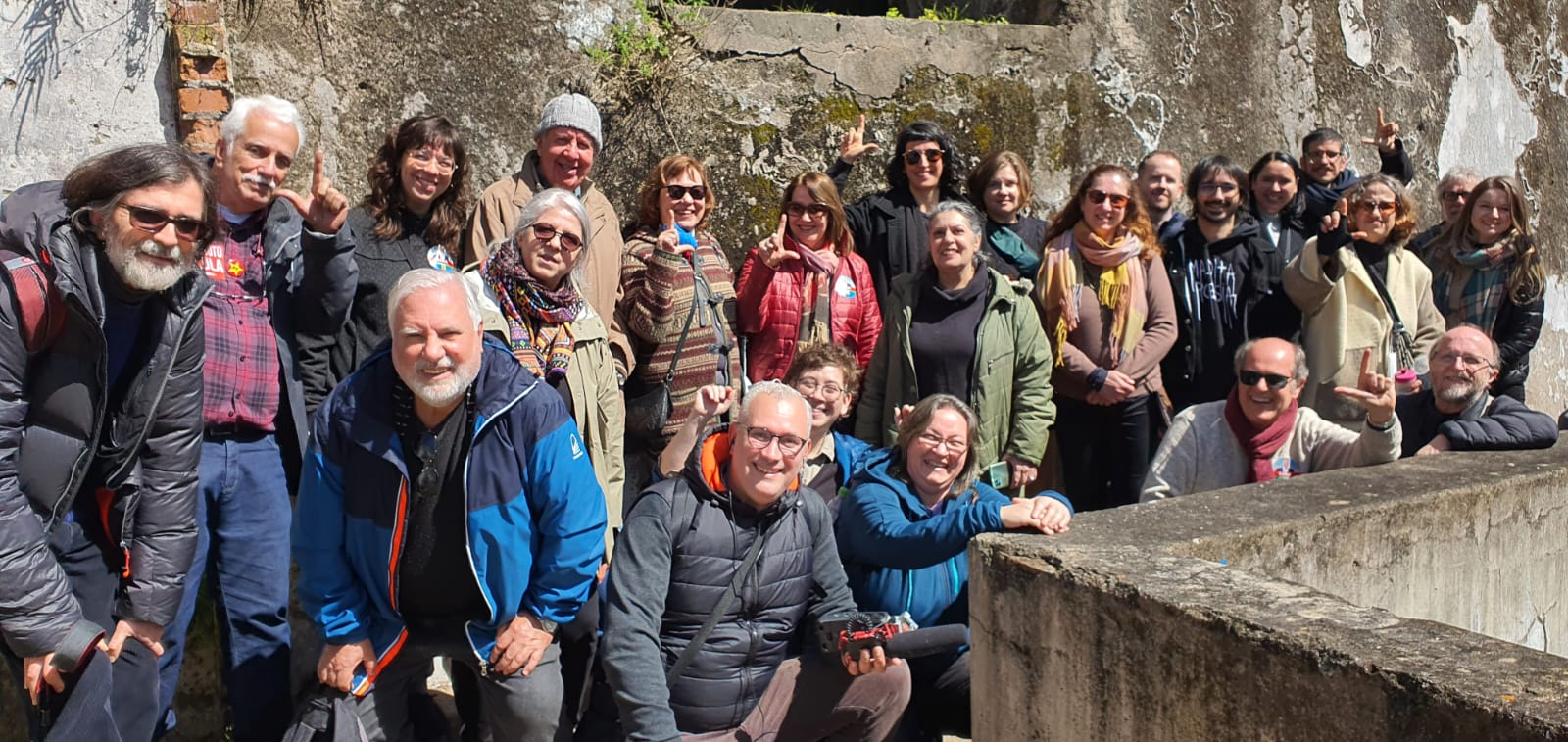 Imagem colorida. Enquadra um grupo de aproximadamente 25 pessoas posando para foto em área externa de ruínas na Ilha do Presídio.